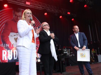 Drei Personen auf Bühne. Frau spricht ins Mikrofon, Landeshauptstadt..jpg