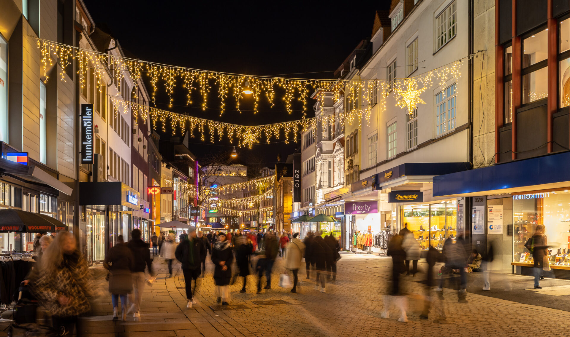 Weihnachtsbeleuchtung über belebter Straße mit Menschen und Geschäften bei Nacht. Dekorative Lichterketten und Sterne schmücken die Szene.