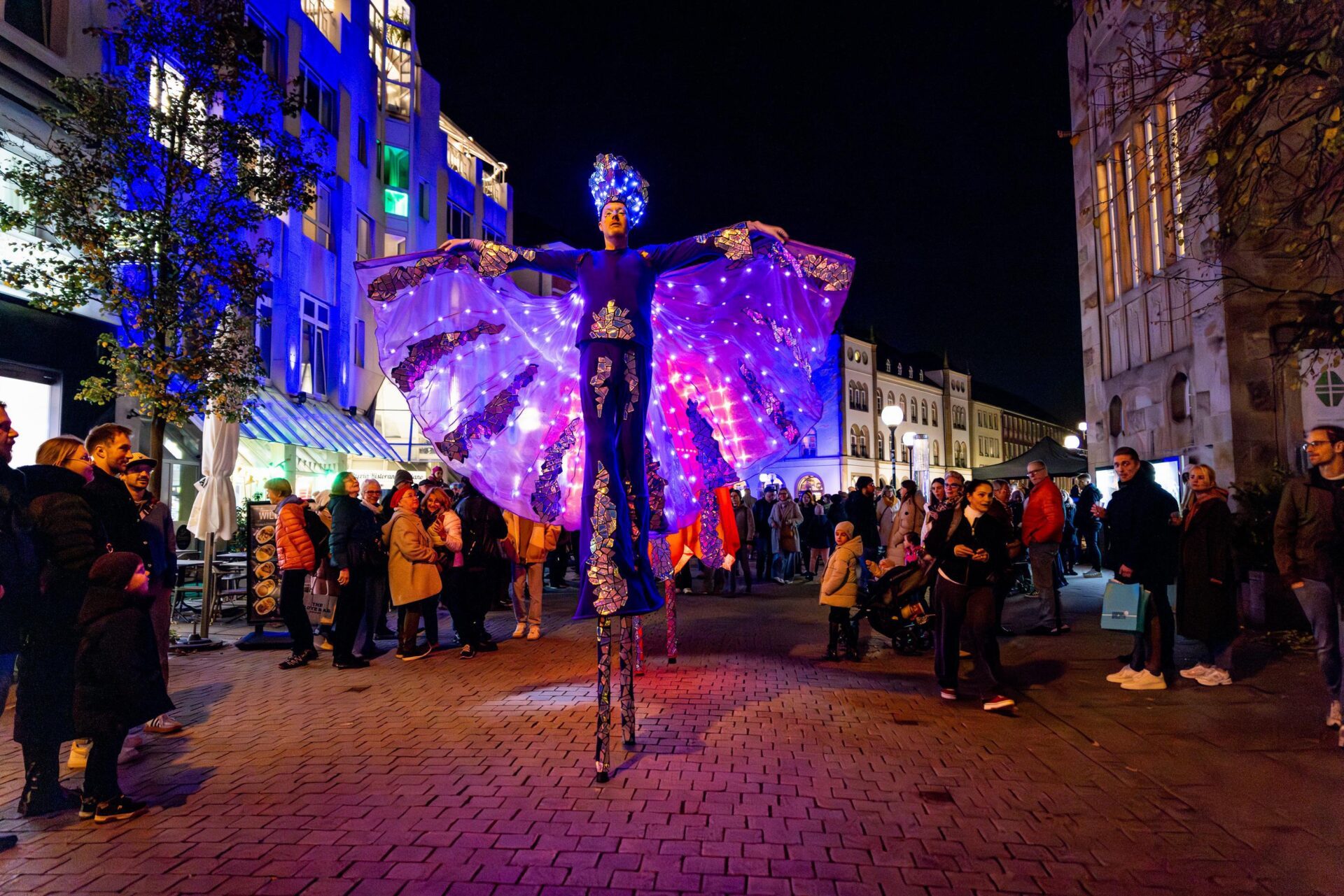 Moonlight Shopping: Stelzenläufer mit Flügeln, beleuchtet. Menschenmenge im Hintergrund.