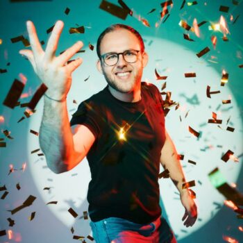 Sven Bensmann Plakat: Smiling man with glasses amid falling confetti, dark shirt, blue jeans, posing for a poster, celebratory mood