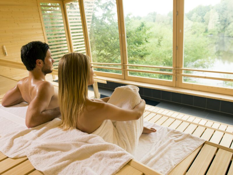 Ein Paar entspannt in einer MeriSauna aus Holz und genießt den Blick auf die Natur durch die Fenster.