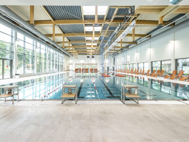 Nettebad 33m Becken: Innenansicht eines Schwimmbads mit Schwimmbecken, Startblöcken und Liegestühlen, helle und moderne Architektur.