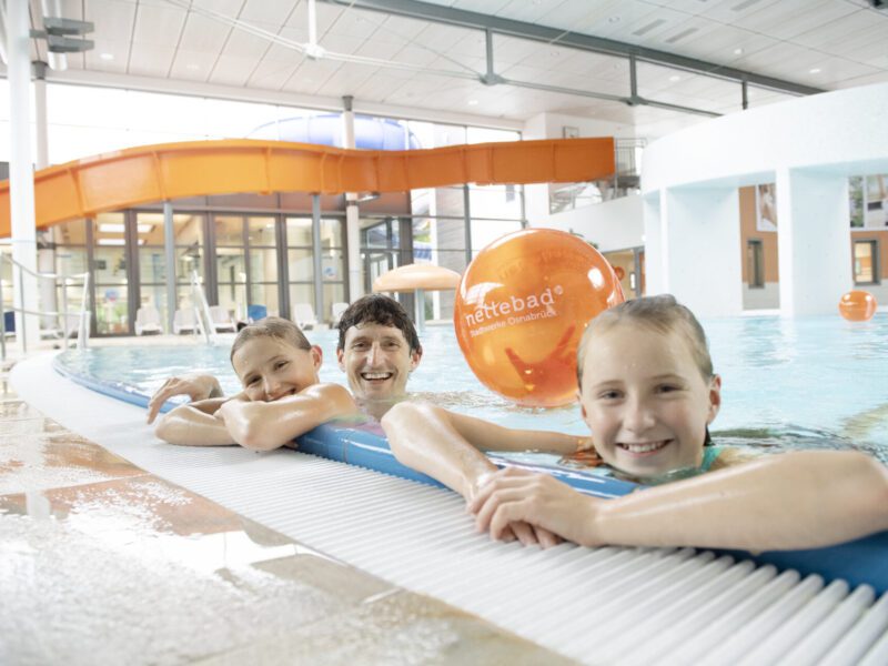 Nettebad Familie: Vater mit zwei Töchtern im Schwimmbad, lachend am Beckenrand. Im Hintergrund eine Rutsche und ein Ball.