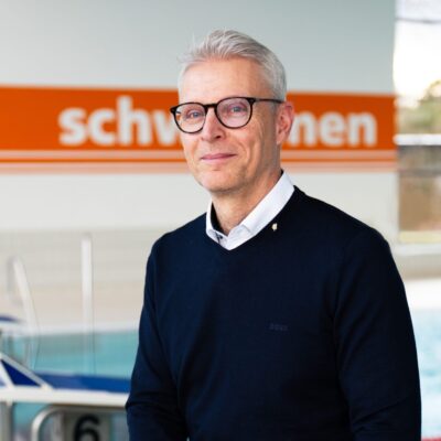 Porträt von Torsten Krier von Stadtwerke vor Schwimmbad Hintergrund. Mann mit Brille und dunklem Pullover.