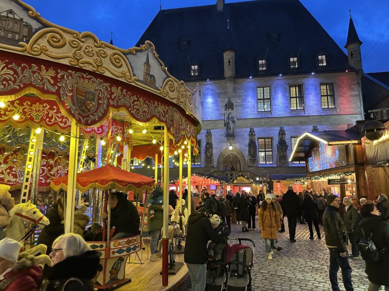 Ein Weihnachtsmarkt-Karussell mit Pferden und Lichtern vor einem historischen Gebäude voller Menschen.