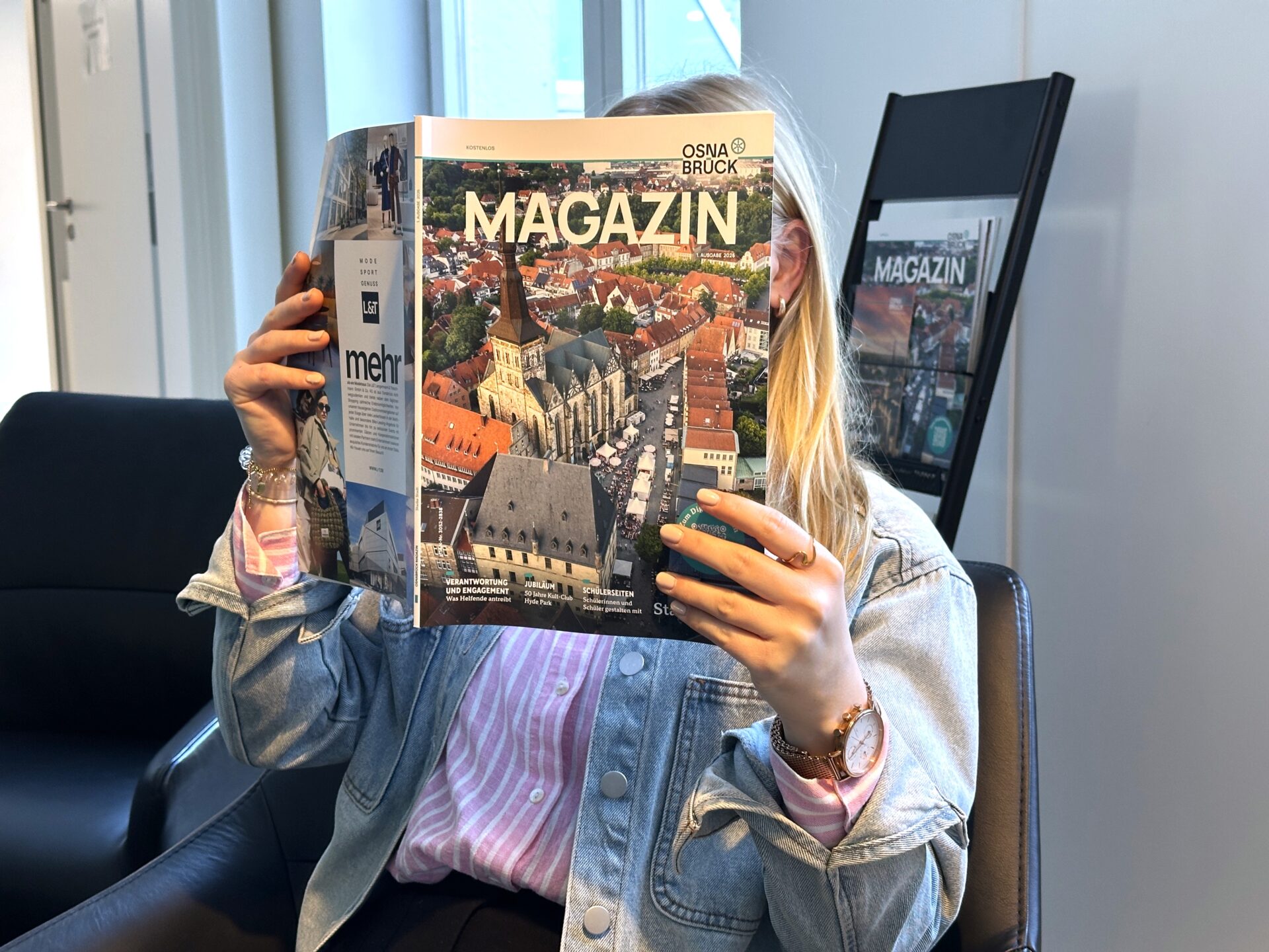 Frau liest OS-Magazin mit Stadtansicht. Das Magazin verdeckt das Gesicht der Leserin. Magazin-Ständer im Hintergrund.