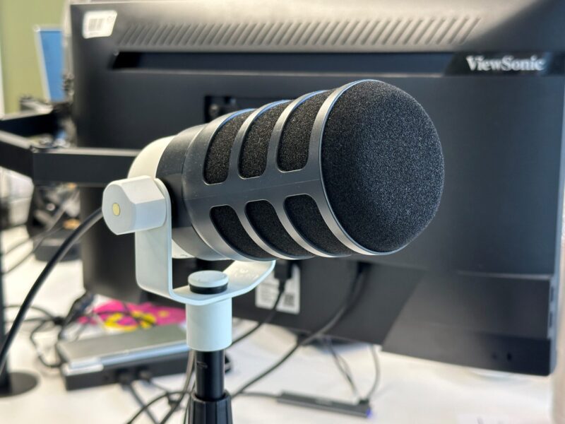 Nahaufnahme eines Podcast-Mikrofons mit schwarzem Windschutz auf einem Ständer vor einem Computerbildschirm.