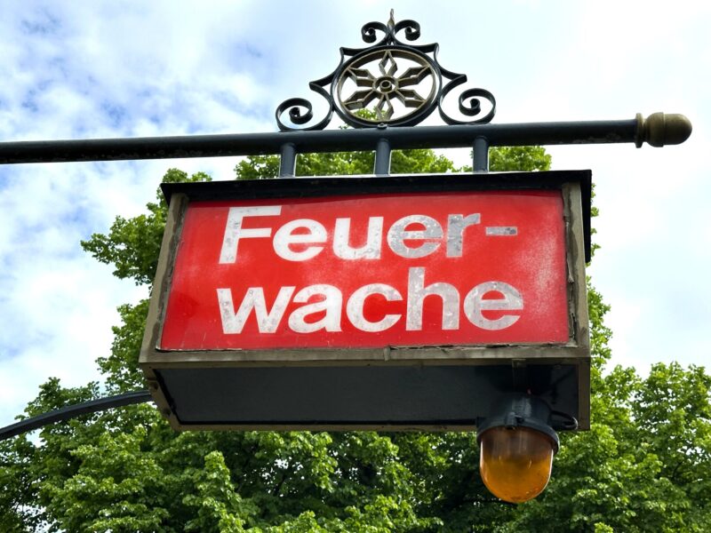 Rotes Schild mit der Aufschrift Feuerwache an einer Halterung mit Lampe vor grünen Bäumen und blauem Himmel