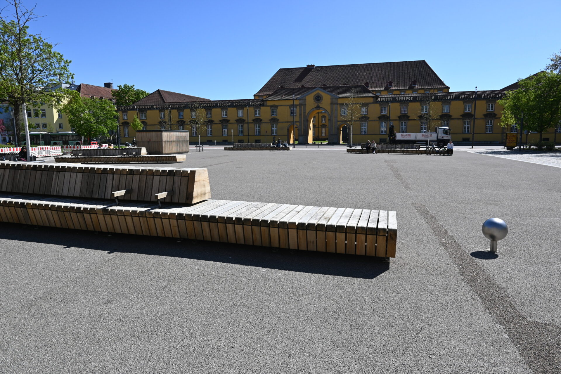Moderner Ledenhof mit großen Holzsitzbänken und gelbem historischen Gebäude im Hintergrund an einem sonnigen Tag