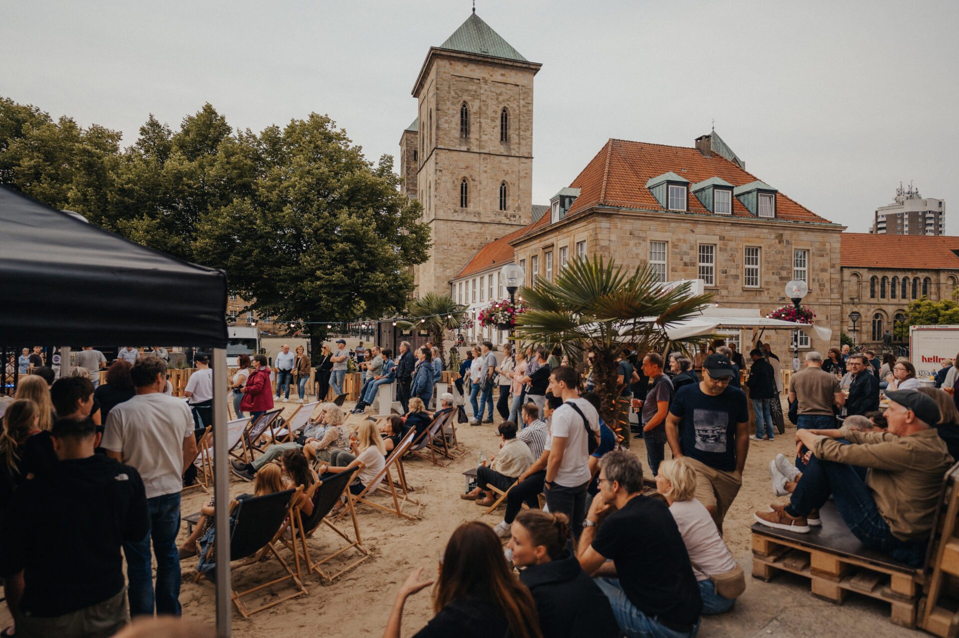 Menschen entspannen auf Liegestühlen in einer sommerlichen Strandbar mit Palmen vor einer historischen Gebäudekulisse