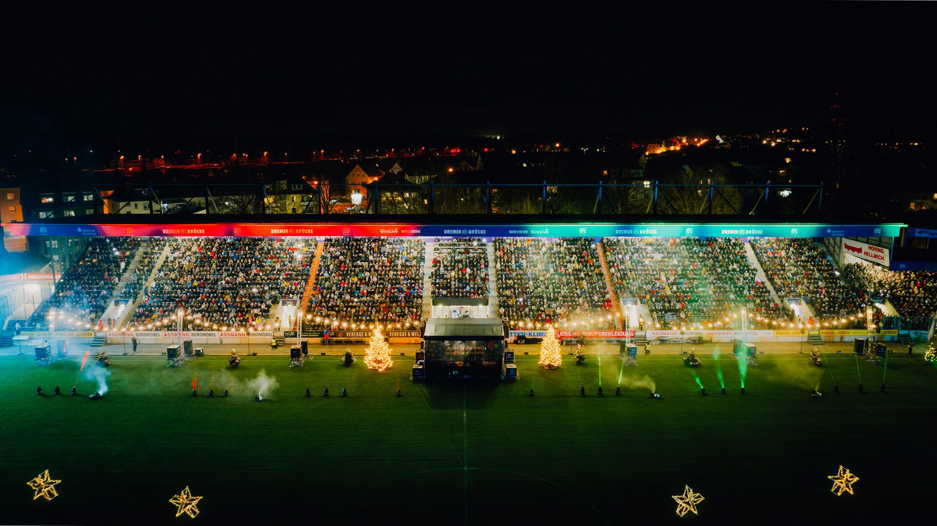 Vollbesetzte Stadiontribüne bei Nacht mit weihnachtlicher Beleuchtung und Tannenbäumen beim gemeinsamen Weihnachtssingen