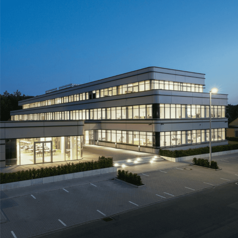 Modernes Bürogebäude bei Dämmerung mit beleuchteten Fensterfronten und vorgelagertem Parkplatz