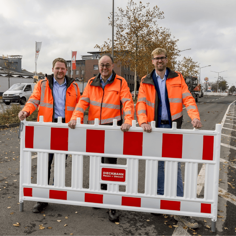 Drei Männer in orangefarbenen Warnwesten stehen hinter einer rot-weißen Absperrschranke mit Dieckmann Logo.