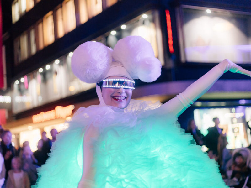 Leuchtende Person in weißem Kostüm mit LED-Brille und Federkopfschmuck bei einer Shopping-Nacht