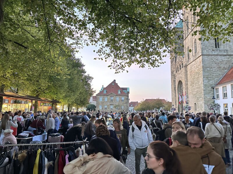 Menschenmenge auf einem nächtlichen Flohmarkt mit Kleiderständen, umgeben von Bäumen und historischen Gebäuden.