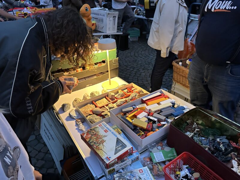 Nachtflohmarkt: Person stöbert bei Abendlicht in Kisten mit Lego-Steinen und Modellbauzubehör.