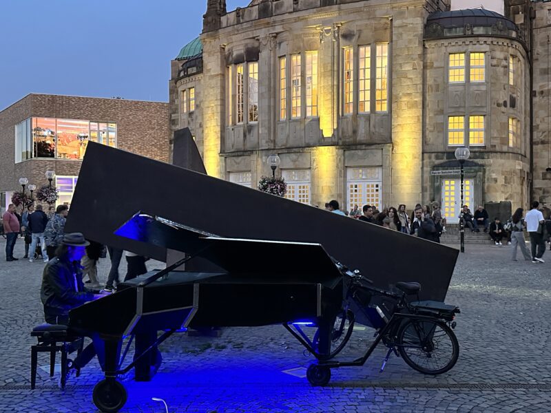 Nachtflohmarkt: Mann spielt auf einem umgebauten, fahrbaren Klavier mit Fahrradanhänger im beleuchteten Innenhof