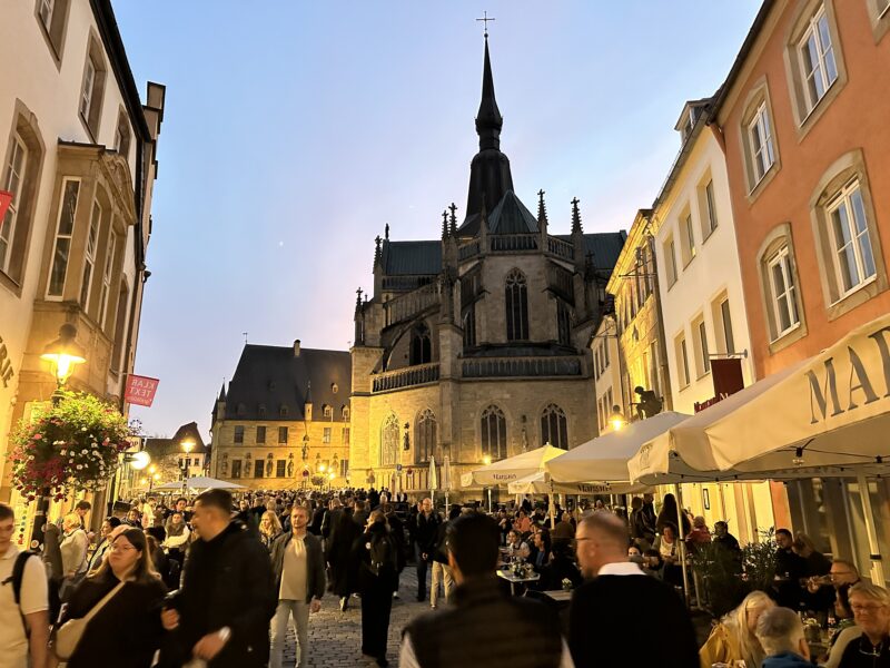 Abendliche Menschenmenge auf einem Nachtflohmarkt in einer historischen Altstadt mit beleuchteter Kirche und gastronomischen Ständen.