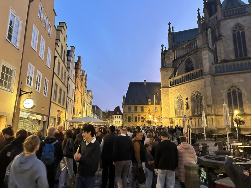 Menschenmenge auf einem nächtlichen Flohmarkt in einer historischen Altstadt mit beleuchteten Fachwerkhäusern und einer großen Kirche im Hintergrund.
